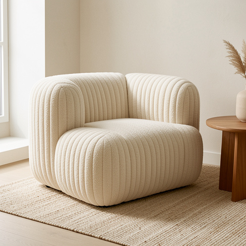 Fauteuil lounge bas en tissu bouclé crème, aux formes arrondies et plissées, installé dans un salon minimaliste lumineux avec une table en bois et un tapis texturé.